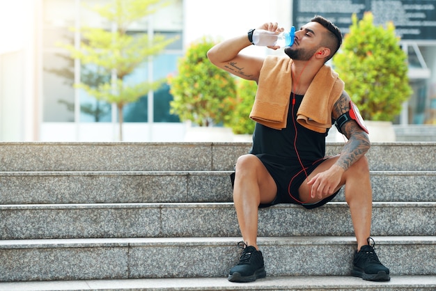 Colocar apuesto joven sentado en los escalones y beber agua después de entrenar al aire libre