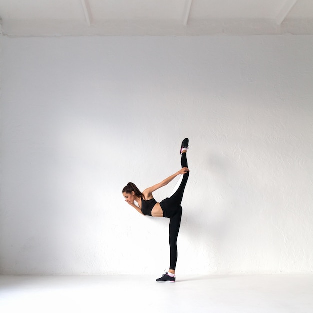 Colocar bastante joven vistiendo top negro y polainas haciendo ejercicios de estiramiento de yoga en el aula de loft de gimnasio blanco de entrenamiento físico. Concepto de deporte estilo de vida saludable. Copie el espacio.