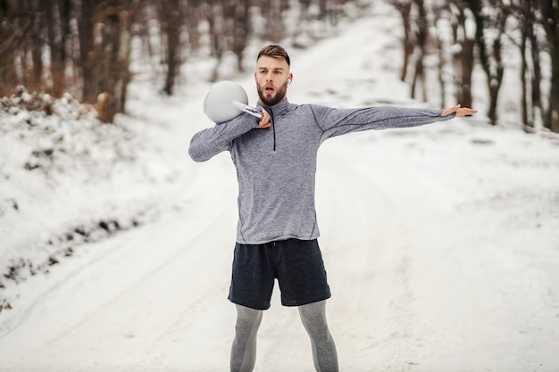 Colocar deportista levantando pesas rusas mientras está de pie en el camino nevado en el bosque. Fitness de invierno, vida sana, culturismo