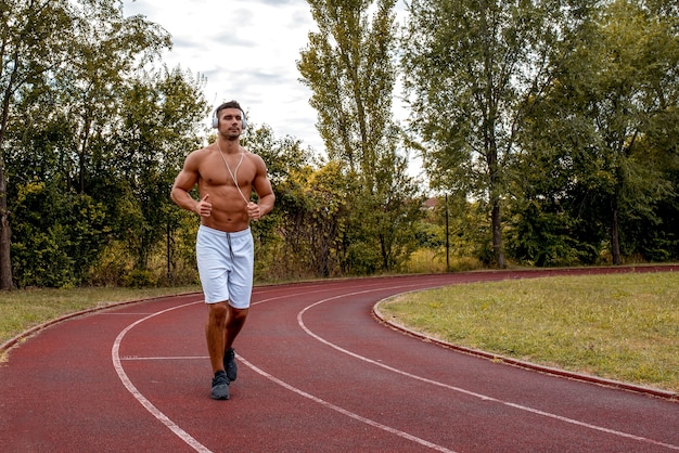 Colocar hombre sin camisa con pantalones cortos blancos y auriculares corriendo en una pista