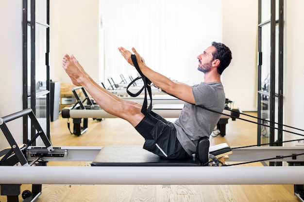 Colocar hombre musculoso haciendo un ejercicio de pilates teaser en un reformador en un gimnasio para fortalecer sus músculos centrales y abdominales en una vista lateral con copyspace