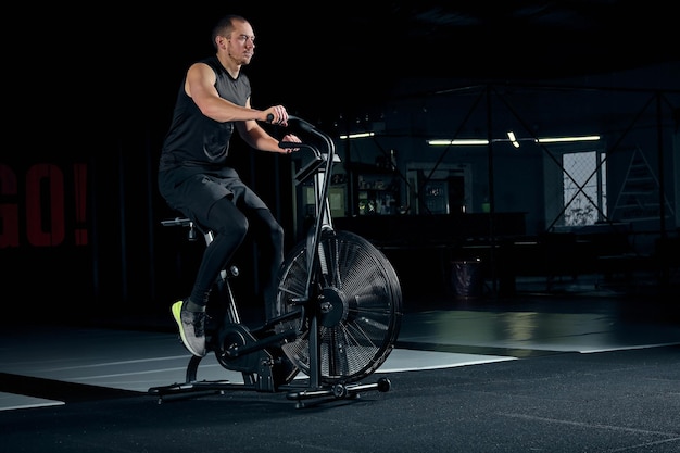 Colocar joven con bicicleta estática en el gimnasio. Hombre de fitness con bicicleta de aire para entrenamiento cardiovascular en el gimnasio de entrenamiento cruzado.