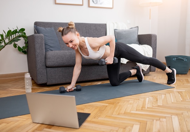 Colocar joven deportista haciendo ejercicio con pesas y viendo videos en la computadora portátil durante el entrenamiento en línea en casa