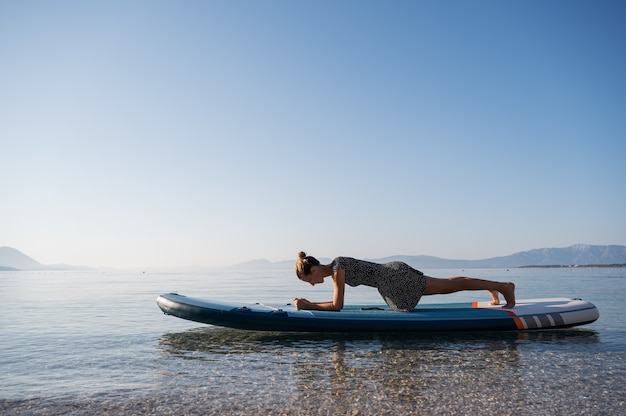 Colocar joven haciendo un ejercicio de fuerza de tabla en una tabla de sup flotando en el agua de mar en calma por la mañana.