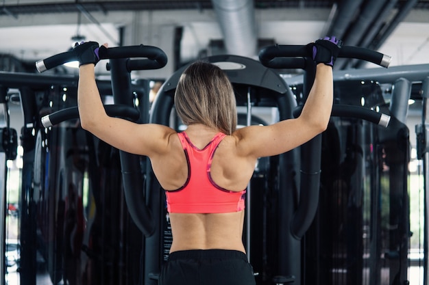 Colocar joven mujer haciendo ejercicio de hombros con máquina de entrenamiento en el gimnasio. Vista desde atrás.