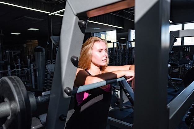 Colocar joven mujer de pie en el gimnasio frente a barra. Retrato de la atleta femenina en una sala de fitness entre un montón de equipos de fitness