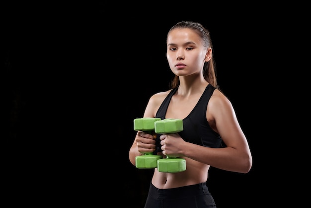 Colocar joven en ropa deportiva sosteniendo pesas por el pecho mientras hace ejercicio contra la pared negra