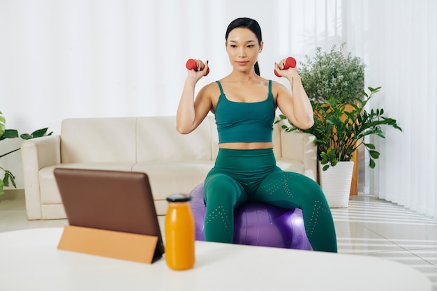 Colocar a una mujer asiática bastante joven sentada en una pelota de fitness, viendo la guía de video en la tableta y haciendo ejercicio con pesas