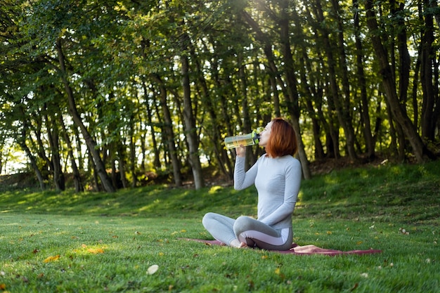 Colocar a la mujer en ropa deportiva bebiendo agua mientras hace ejercicio, haciendo yoga en el parque en un día soleado de otoño.