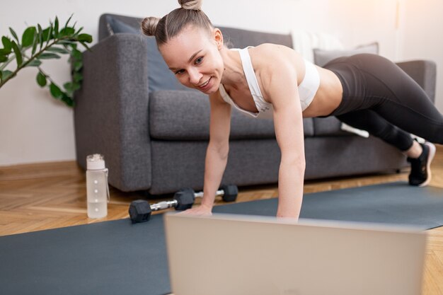 Colocar a la mujer en ropa deportiva sonriendo y viendo videos en la computadora portátil mientras hace ejercicio de tabla en la alfombra durante la cuarentena en casa