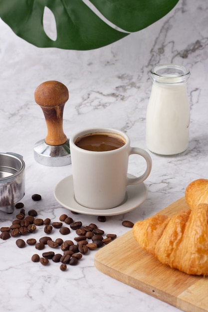 Se colocó una taza de café con leche junto a una botella de leche y un croissant en una tabla de cortar, granos de café y molinillos en un piso de mármol.