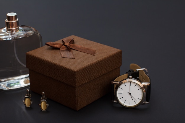 Colonia para hombre, caja regalo marrón, gemelos y reloj con correa de piel negra sobre fondo negro. Complementos para hombre.