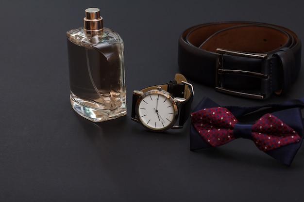 Colonia para hombre, reloj con correa de piel negra, pajarita y cinturón de piel con hebilla metálica sobre fondo negro. Complementos para hombre.