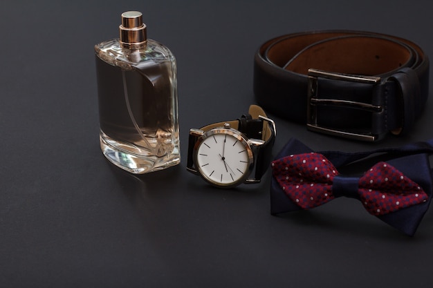 Colônia para homem, relógio com pulseira de couro preta, gravata borboleta e cinto de couro com fivela de metal em fundo preto. Acessórios para homem.