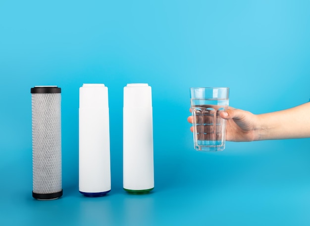 Coloque tres cartuchos para filtro de agua con un vaso de agua transparente sosteniendo la mano sobre fondo azul