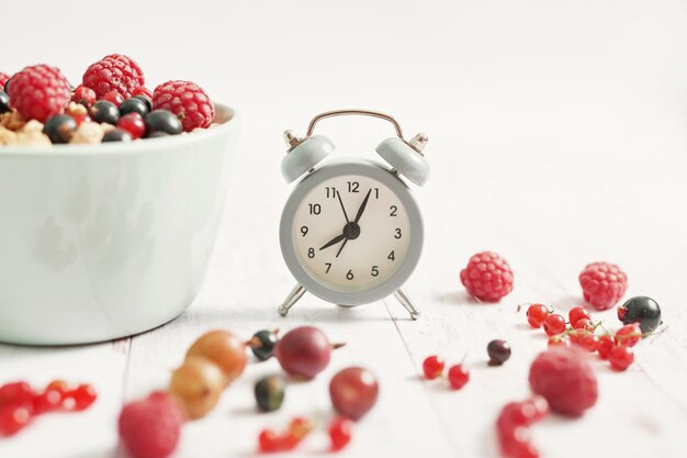 Coloridas bayas frescas con escamas en una taza blanca y reloj despertador