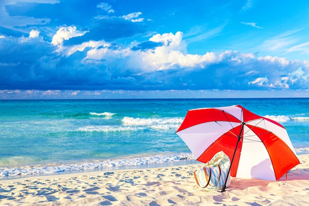 Colorido paraguas rojo y blanco con bolsa de playa en la playa del océano contra el hermoso cielo azul y las nubes. Relajación, fondo idílico de vacaciones.