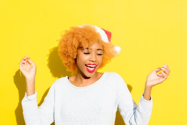 Colorido retrato de una linda mujer africana con sombrero de Navidad sobre fondo amarillo. Concepto de vacaciones de invierno