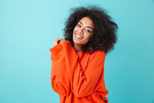 Colorido retrato de mujer afroamericana en camisa roja con peinado afro mirando con una sonrisa presionando sus brazos contra el pecho, aislado sobre la pared azul