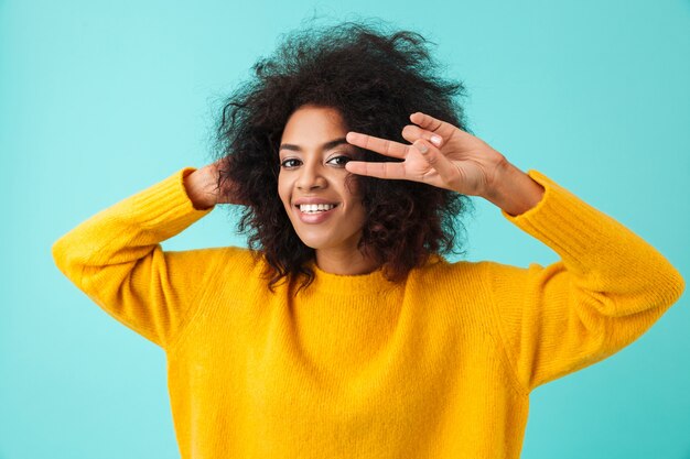 Colorido retrato de mujer increíble con cabello peludo sonriendo y mostrando el símbolo de la victoria con los dedos, aislado sobre la pared azul