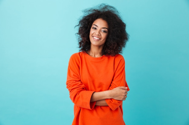 Colorido retrato de mujer increíble en camisa roja con peinado afro mirando con sonrisa, aislado sobre la pared azul