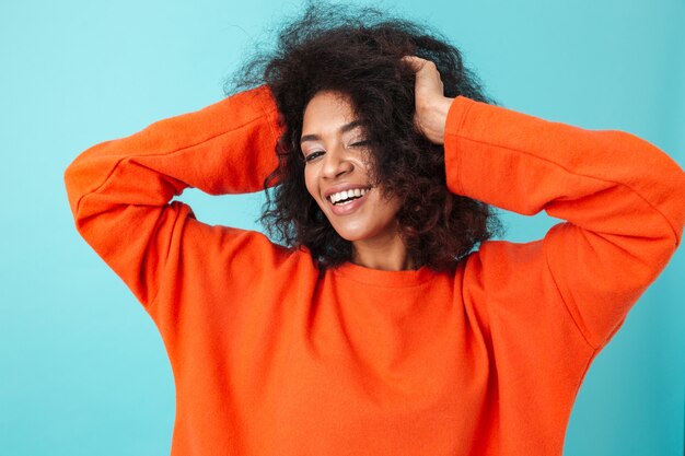 Colorido retrato de mujer sonriente en camisa roja posando y tocando su cabello oscuro y rizado, aislado sobre la pared azul