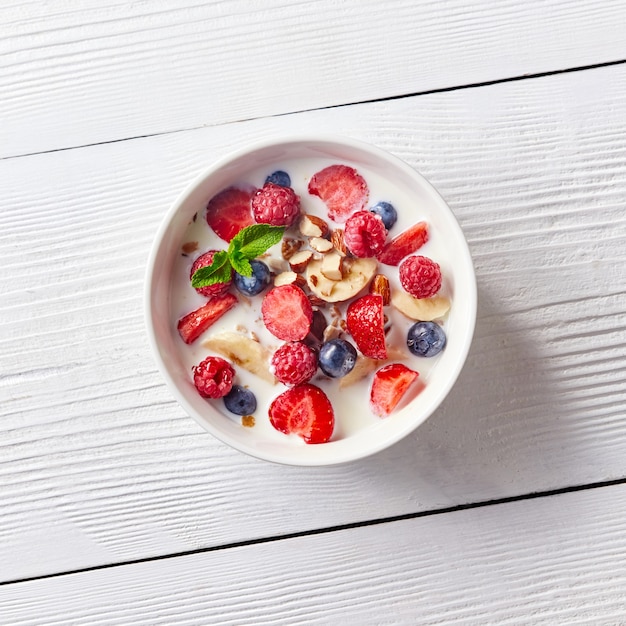 Colorido y sabroso desayuno preparado de muesli de maíz, bayas, leche, almendras, rodajas de plátano en un recipiente sobre una mesa de madera blanca con espacio de copia. Endecha plana.