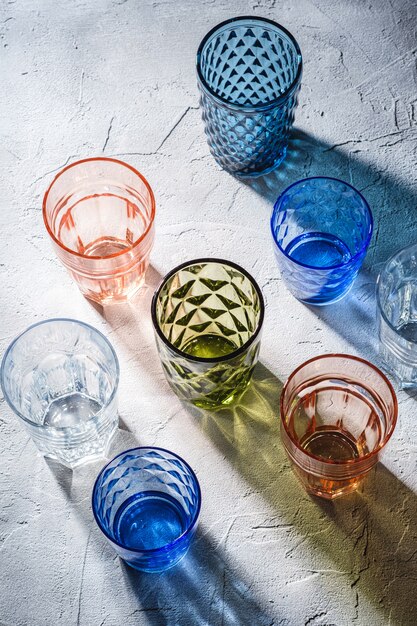 Colorido vaso de bebida facetado y geométrico, grupo de vasos vacíos verdes, rojos, azules y transparentes para bebidas en la mesa de hormigón de piedra, vista de ángulo