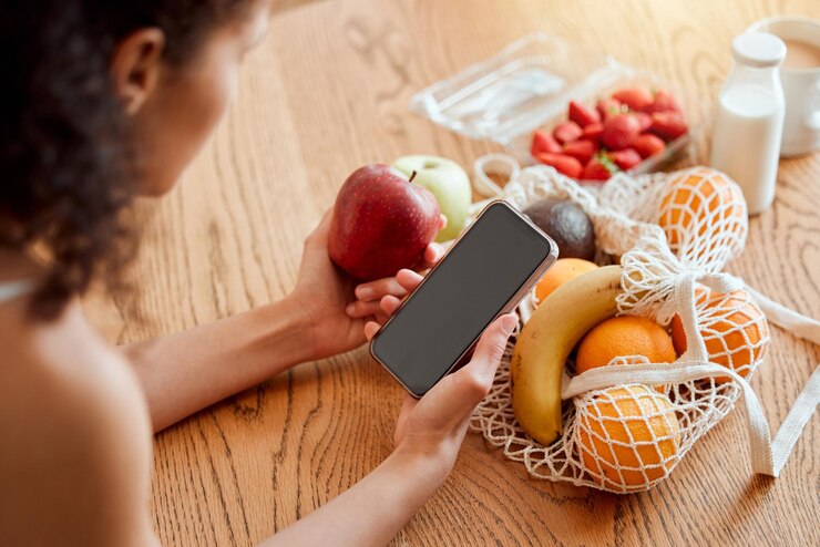 Foto coloridos comestibles de frutas orgánicas saludables y nutricionales en la mesa junto a la mujer que sostiene el teléfono y busca en línea recetas de batidos jovencita comiendo manzana orgánica fresca para una dieta vegana de bienestar