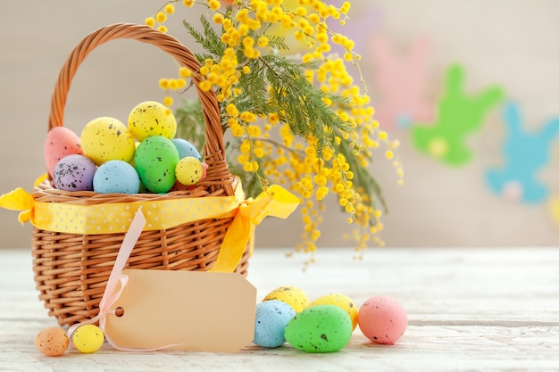 Coloridos huevos de pascua en una canasta sobre un fondo de madera blanca, decorada con una ramita de flores de mimosa. Concepto de vacaciones de primavera.