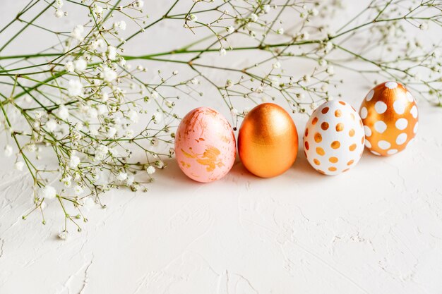 Coloridos huevos de Pascua pintados en oro, blanco y rosa sobre fondo blanco decorado con flores de gypsophila.