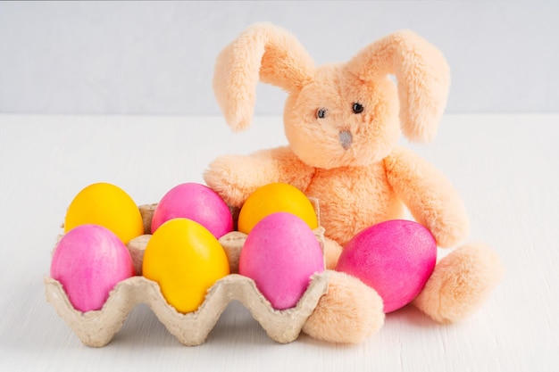 Coloridos huevos de Pascua rosados y amarillos hervidos con juguete de conejito esponjoso sobre fondo blanco de madera