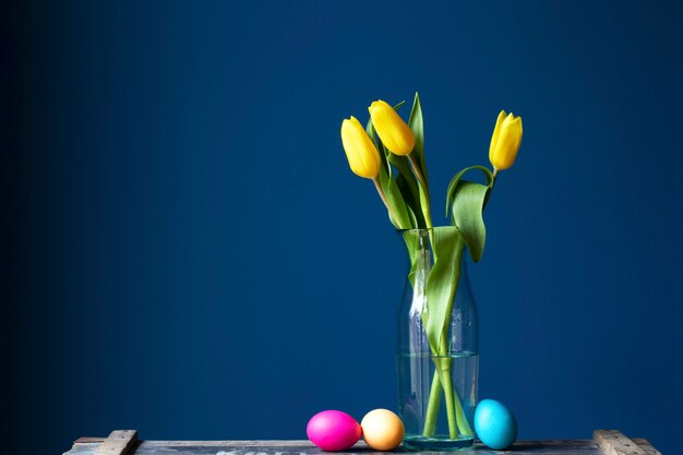 Coloridos huevos de pascua teñidos en una textura de estante de madera vintage cerca de la pared azul con tulipanes amarillos frescos en florero de vidrio pasteles suaves y hojas verdes de diseño moderno