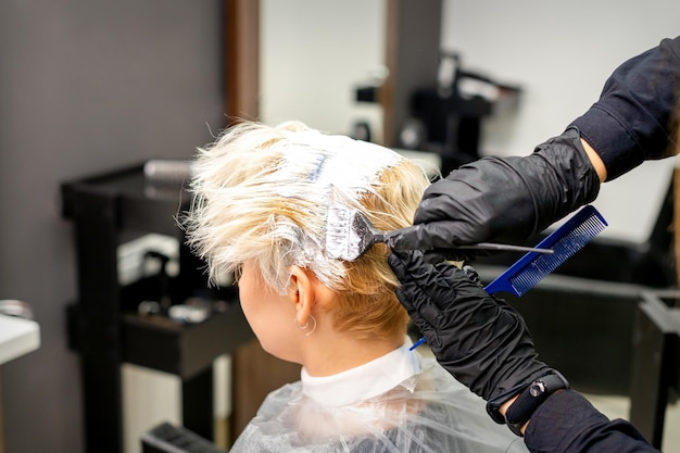 Colorir o cabelo branco com tintura de cabelo e escova pelas mãos do cabeleireiro para a jovem loira caucasiana em um salão de cabeleireiro close-up