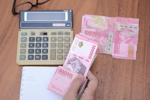 Colseup Die Hand einer Frau, die einige indonesische Rupiah hält, ein Taschenrechner, ein Notizbuch und eine Brille sind in der Nähe