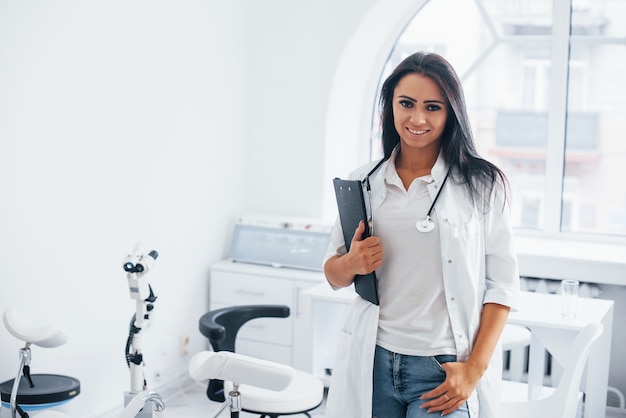 Com bloco de notas nas mãos. Morena médica fica em um escritório moderno e olha para a câmera.