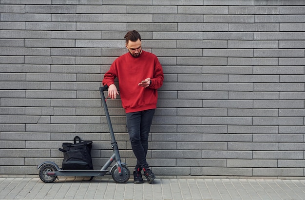 Com bolsa preta um jovem bonito em roupas casuais em pé com uma scooter elétrica ao ar livre no dia ensolarado