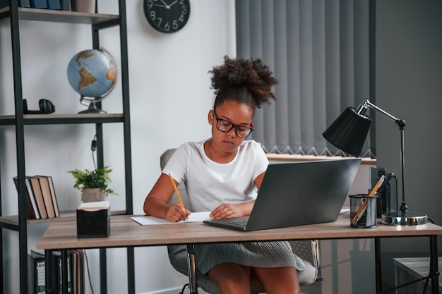 Com caneta, uma linda estudante afro-americana está sentada ao lado do laptop