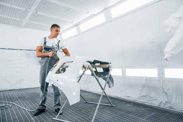 Com pistola de pintura, reparador de automóveis caucasiano em uniforme trabalha na garagem