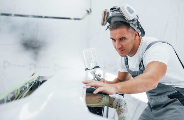 Com pistola de pintura, reparador de automóveis caucasiano em uniforme trabalha na garagem