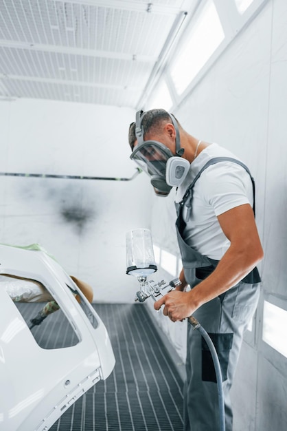 Foto com pistola de pintura, reparador de automóveis caucasiano em uniforme trabalha na garagem