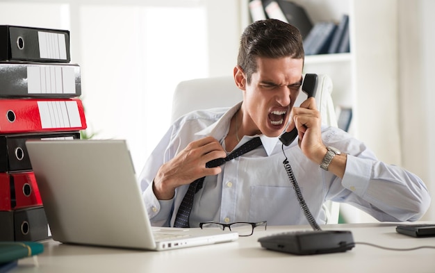 Com raiva jovem empresário sentado no escritório e gritando ao telefone.