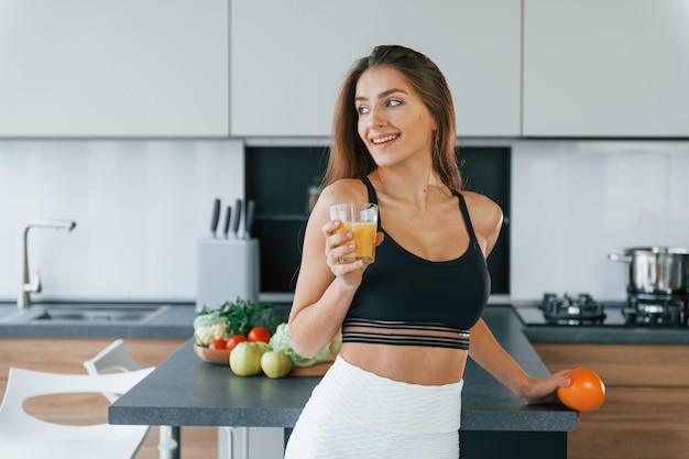 Com suco de laranja fresco a jovem europeia está dentro de casa na cozinha com comida saudável