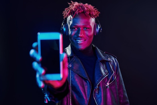 Com telefone e fones de ouvido Iluminação de neon futurista Jovem afro-americano no estúdio