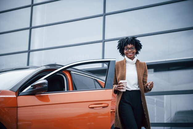 Com um copo de bebida. Jovem mulher afro-americana de óculos fica ao ar livre perto de um carro moderno.