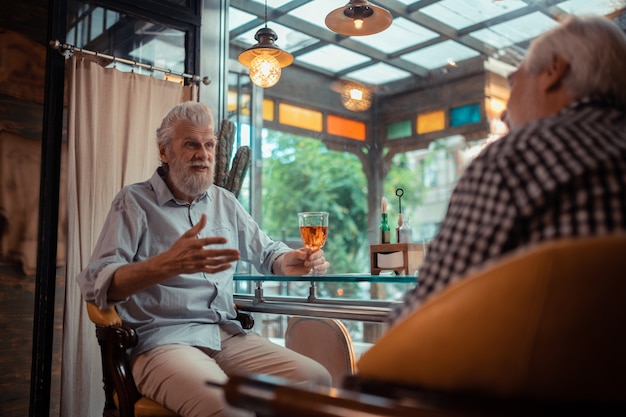 Com um velho amigo. Homem barbudo de cabelos grisalhos sentado em um bar bebendo álcool com um velho amigo