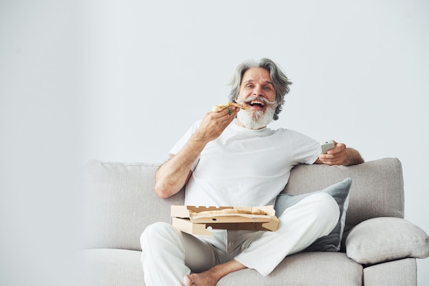 Com uma deliciosa pizza, assiste a um programa de televisão, um homem moderno e elegante, com cabelos cinzentos e barba, dentro de casa.