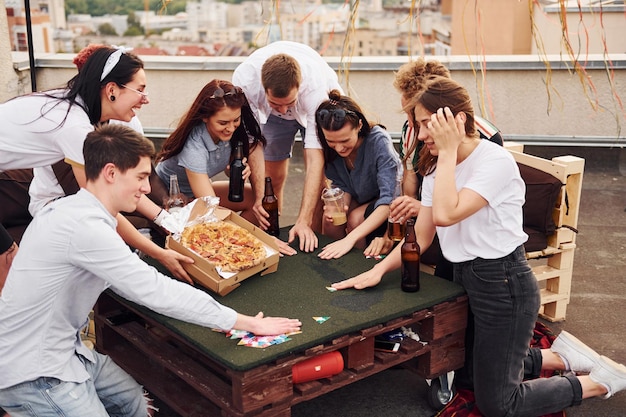 Com uma deliciosa pizza grupo de jovens em roupas casuais fazem uma festa no telhado juntos durante o dia