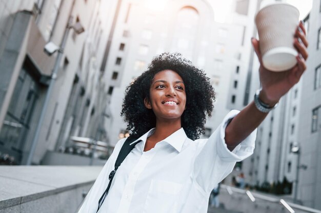 Com uma xícara de bebida, a mulher afro-americana em boas roupas está ao ar livre na cidade durante o dia