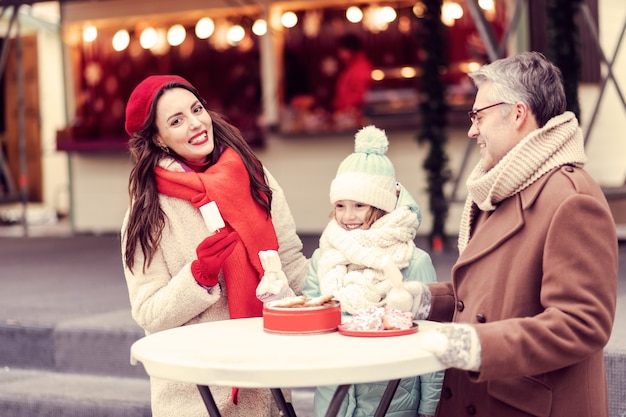 Comamos. Chica alegre manteniendo una sonrisa en su rostro mientras está de pie entre sus padres